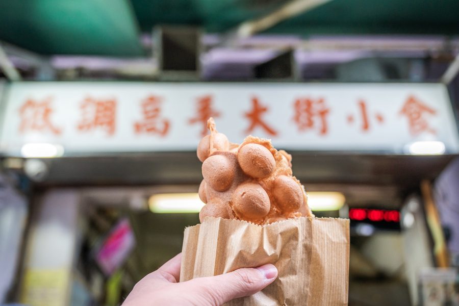 A hand holding a paper bag of egg waffles in front of Master Low-Key Food Shop.  