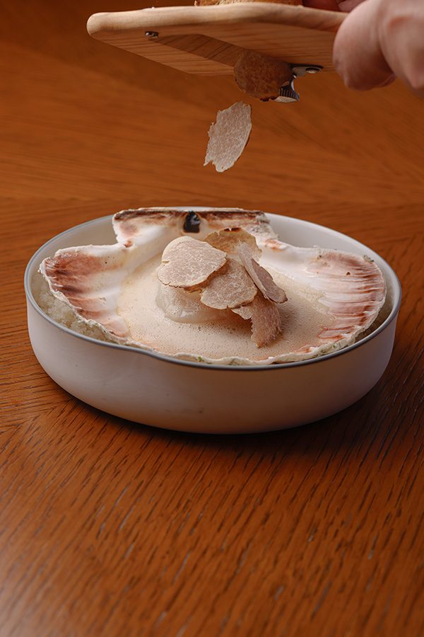 A close up of a person garnishing a white dish of Normandy scallop with white Alba truffle slices, served at Amber, Mandarin Oriental in Central. 