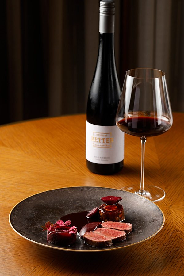A black plate of roe deer loin, neck and foie gras a la royal with beetroot, mas a miel and plum with a glass and bottle of Obsthof Retter red wine on a wooden table, served at Amber, Mandarin Oriental in Central. 