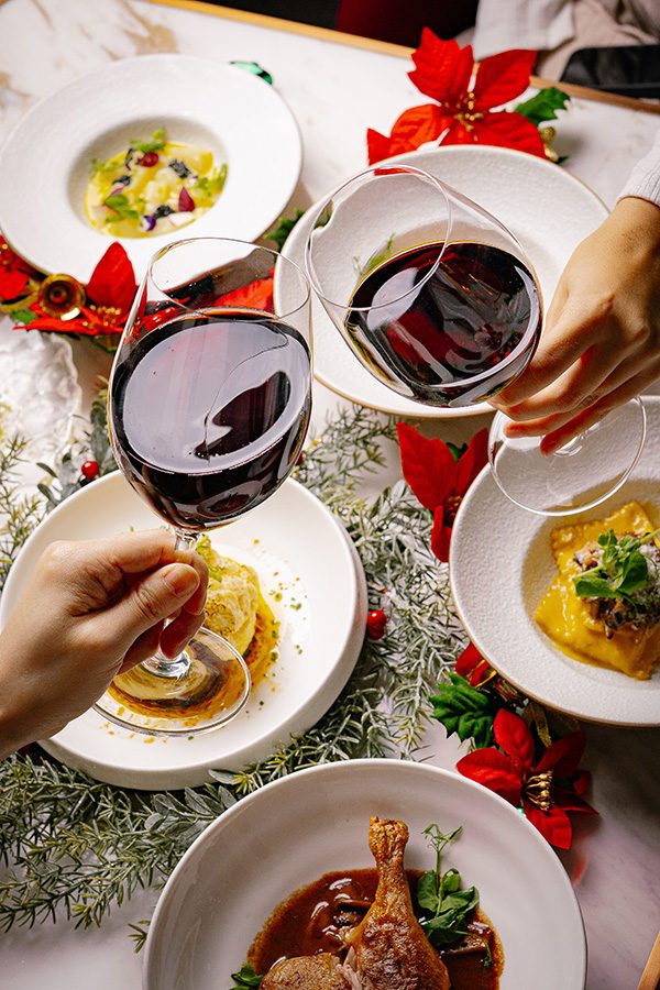 A closeup of two people clinking glasses of red wine over a dining table decorated with Christmas decoration and laid with a spread of various festive dishes served at Bianco & Rosso in Central.