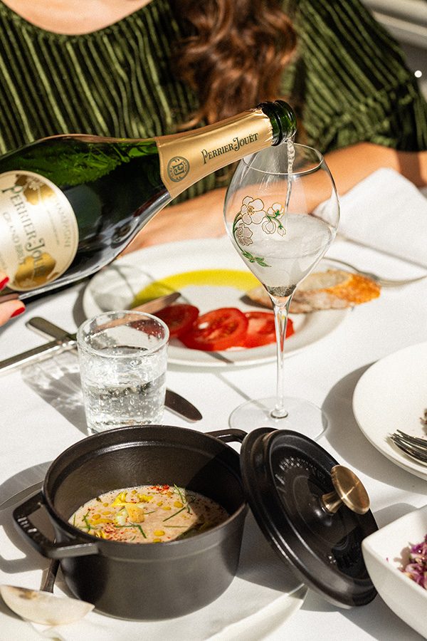  A closeup of a person serving a bottle of perrier-jouët grand brut champagne in a wine glass for a woman at the dining table filled with food at La Petite Maison in Central. 