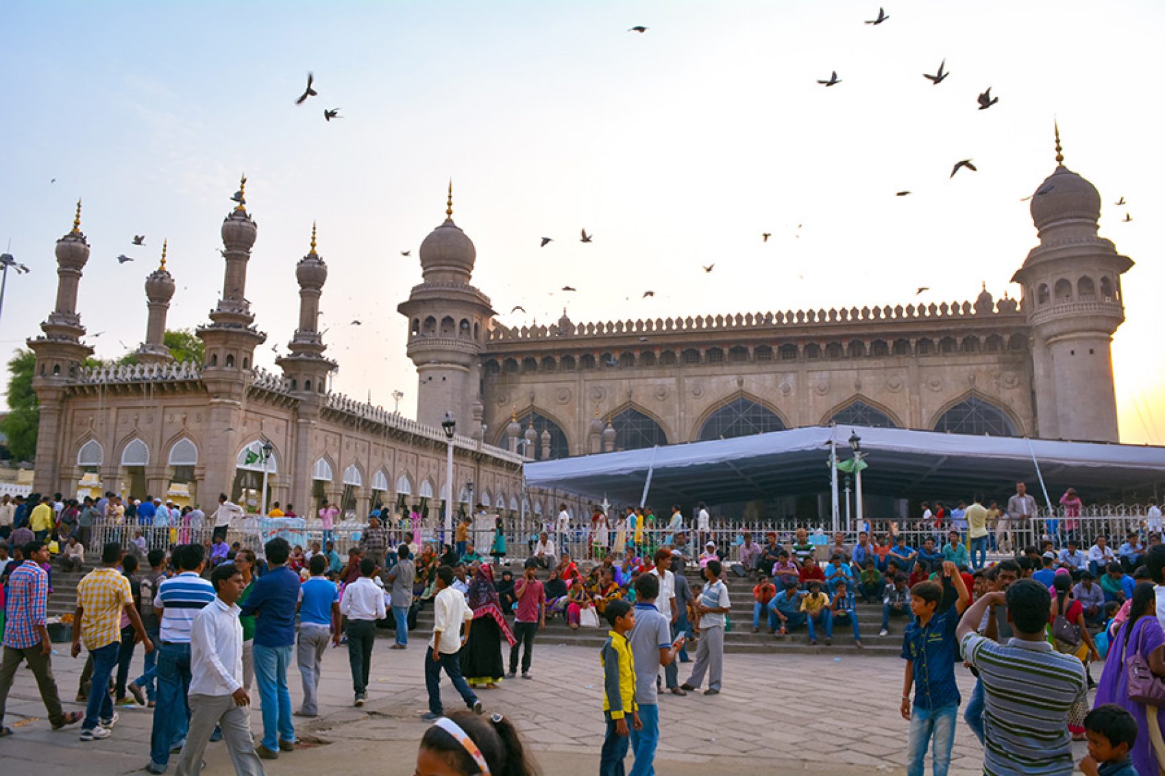 A crowd standing outside of the Mecca Masjid mosque at daytime as birds fly above, in Hyderabad, India.