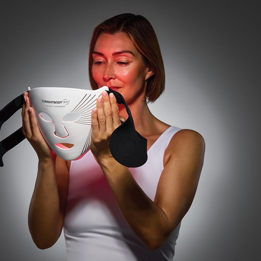 A woman in a white top holding Currentbody Skin’s LED Light Therapy Mask to her face.