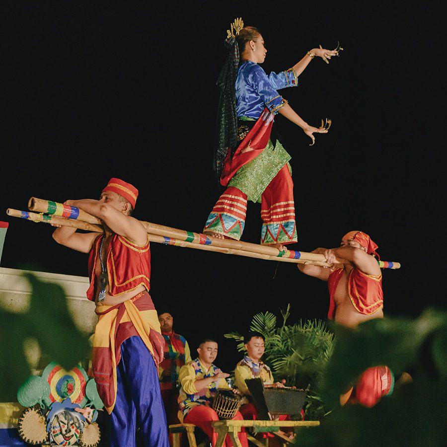 Two male performers at the Shangri-La Mactan’s Kasadya Night in Cebu hold a pair of bamboo poles on their shoulders. A female performer stands on the poles. 
