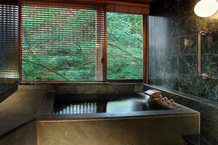 The bath at a hot spring room at Taipei Radium Kagaya International Hotel, designed with natural stone and a window facing green foliage. 