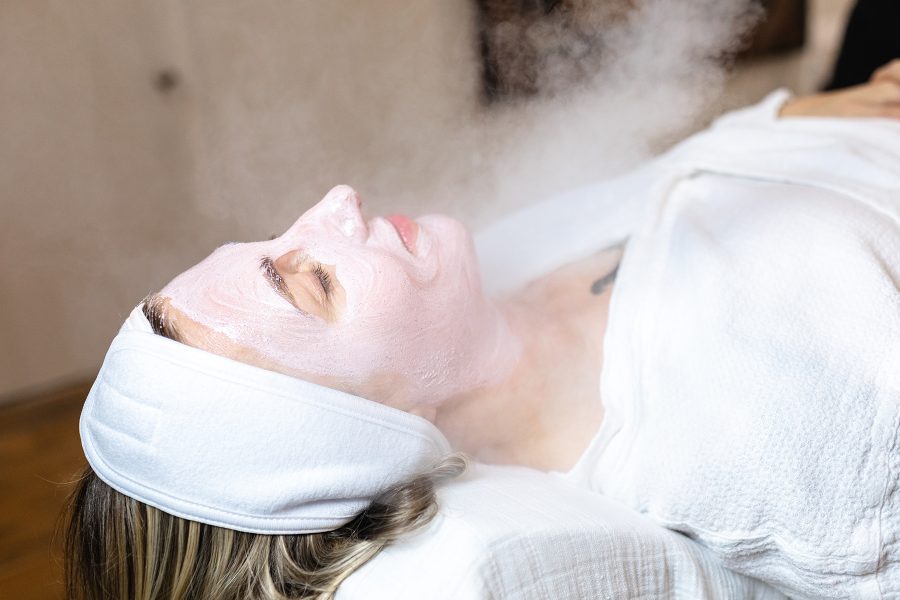 A woman lies down with her eyes closed and a mask on her face as steam billows upward in the background. 