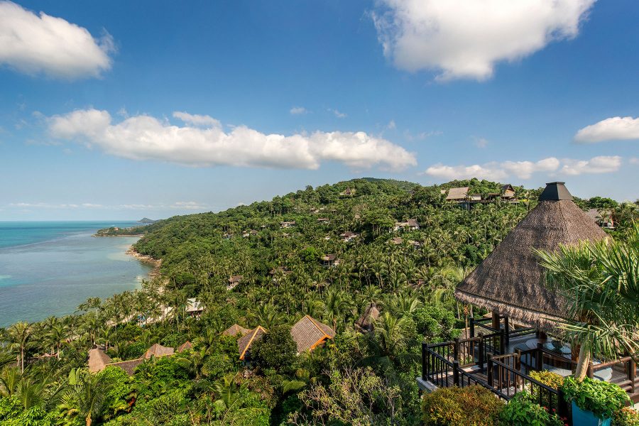 Four Seasons at Koh Samui from above features a sweeping view of rolling green hills sloping into the sea as a brown hut stands in the foreground. 