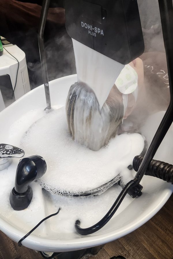 A woman lies down with her head over a sink as a fountain of water pours over her hair and into the soap-filled sink. 