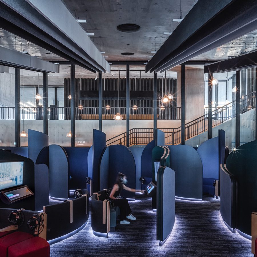 Interior of M+’s Mediatheque. A woman is sitting in one of the blue Viewing Booths with headphones on, reaching towards a touchscreen. 