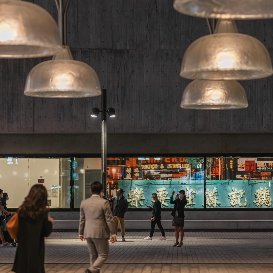 People walking in front of M+, with a view of the museum’s neon sign exhibition through the window.