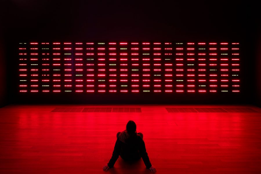 A person sitting on the floor in front of Miyajima Tatsuo’s Region No. 43701–No. 43900, a dark wall with red lights. 