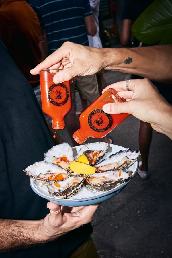 Two hands holding two bottles of Flagrant Sauce over a plate of oysters. 