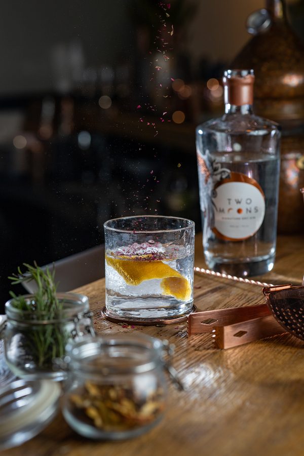 A wooden counter with a glass with lemon slices and a bottle of gin from Two Moons Distillery.  