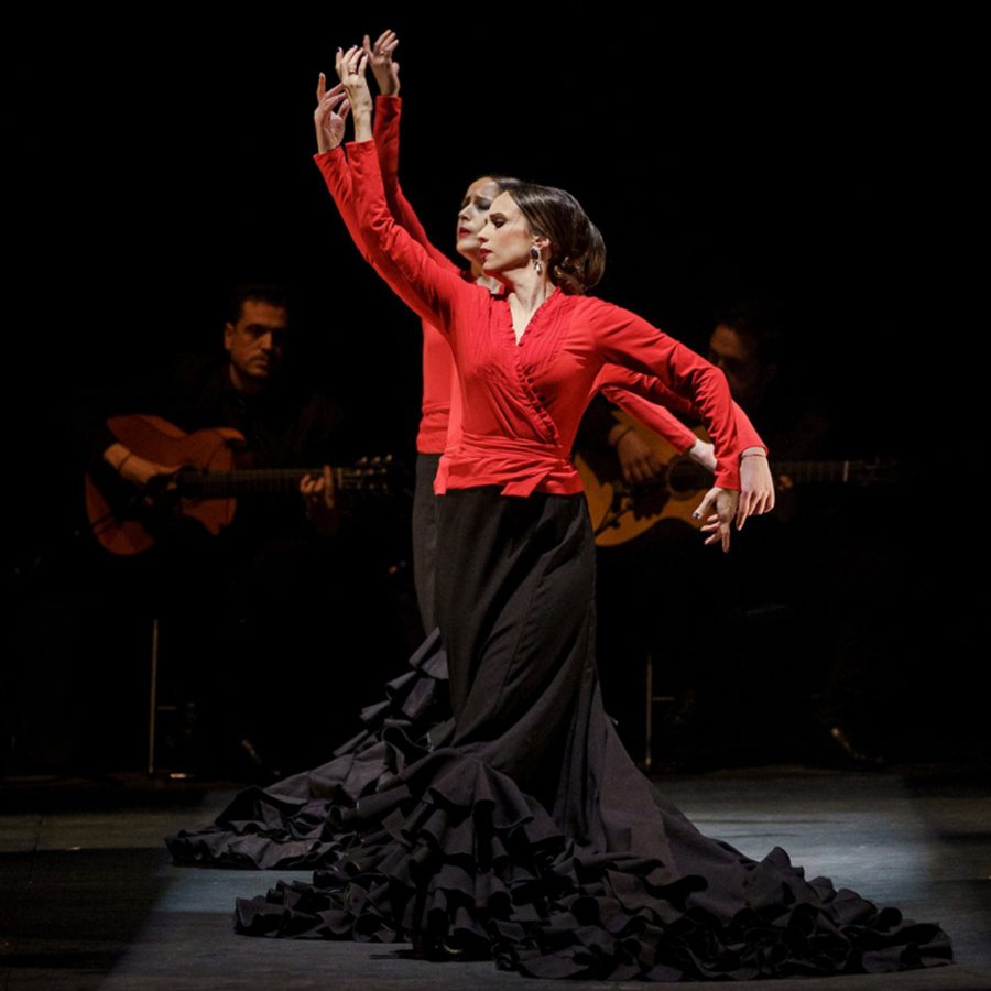 Dancers in red and black Flamenco pose on a dim stage while behind them sit two guitarists, during a performance of Carmen. 