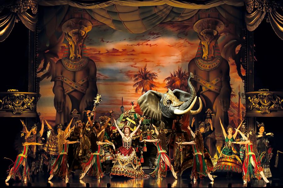 During a production of the musical The Phantom of the Opera, performers in colourful costumes stand in various poses on a stage alongside a mechanical elephant. 
