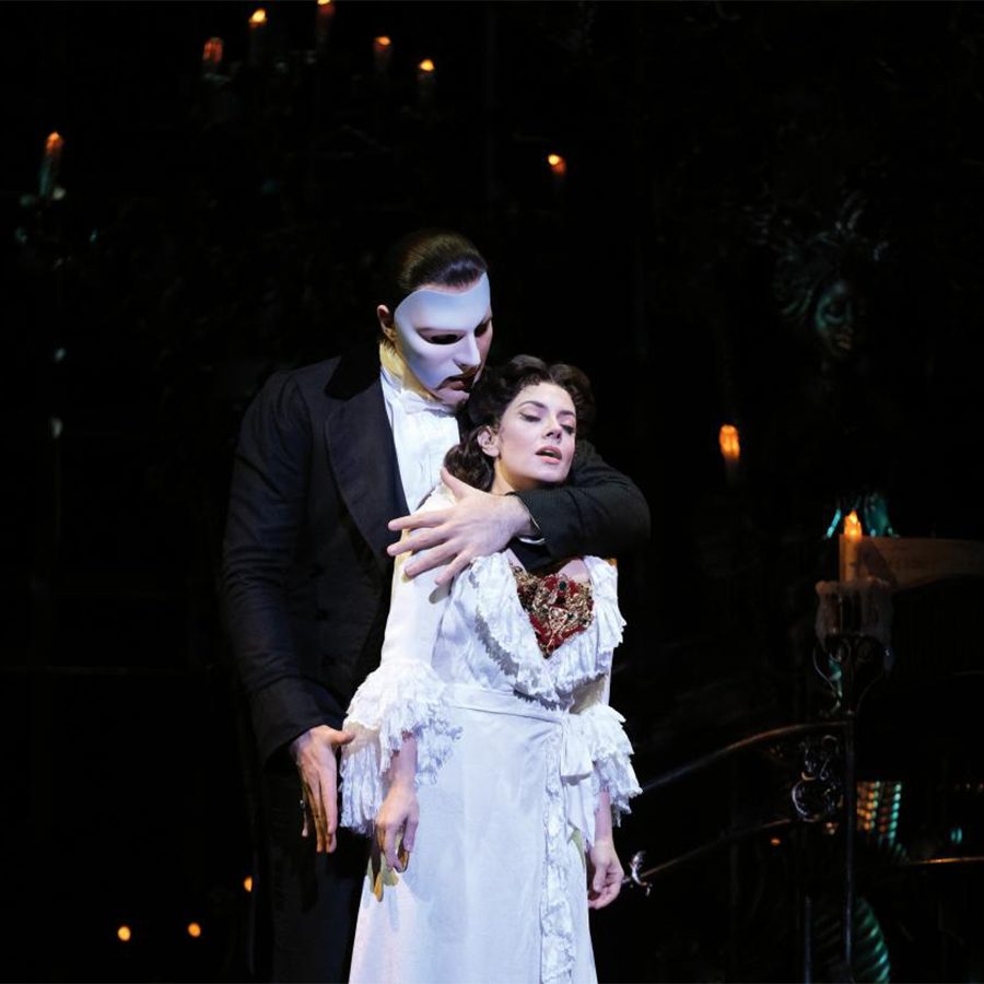 A man with a mask over his face and a woman in a white dress embrace on a softly lit stage, during a production of the musical The Phantom of the Opera. 