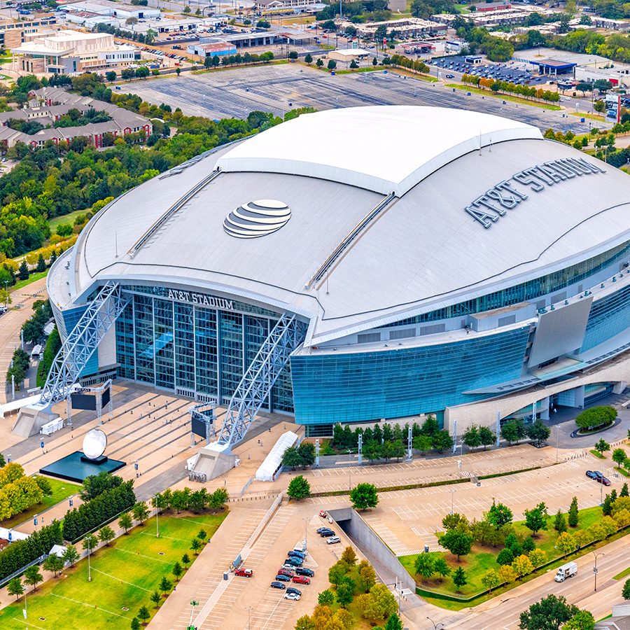 Aerial view of AT&T stadium, standing among green spaces and low suburban buildings, where you can watch a Dallas Cowboys football game.