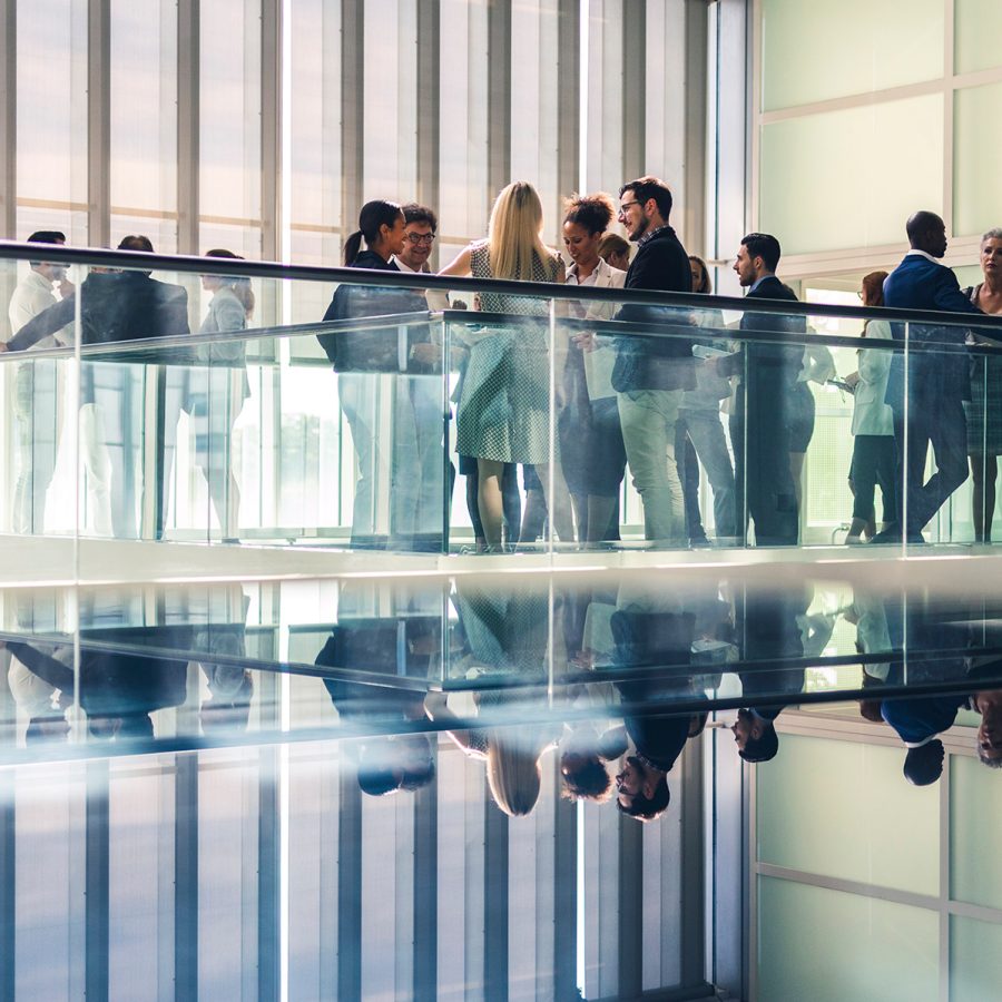 Groups of people dressed in formalwear mingle atop a glass-barricaded walkway inside a building.