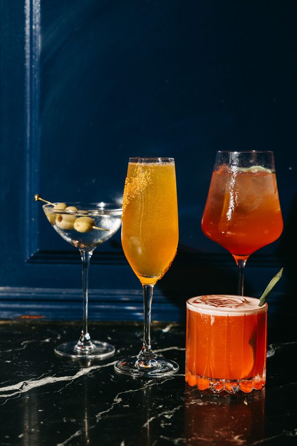A few cocktails and drinks are arranged atop a black marble table in front of a blue wall at The French Room at The Adolphus Hotel. 