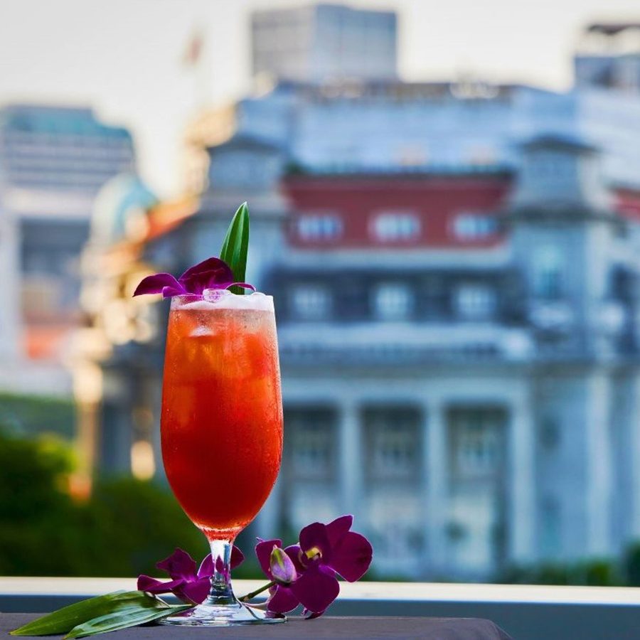 A red cocktail sprinkled with purple flowers, stands against the backdrop of The Fullerton Hotel Singapore.