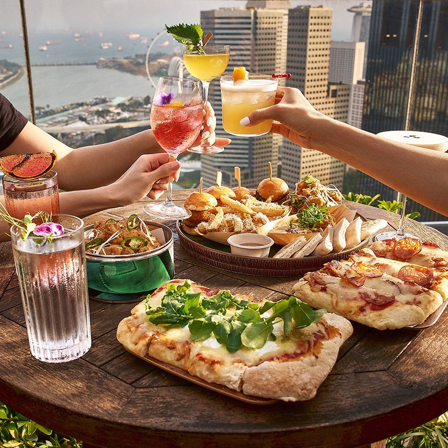 Diners clink cocktail glasses over a meal of flatbread, pizzas and burger sliders as Singapore’s skyscrapers stand tall in the background.