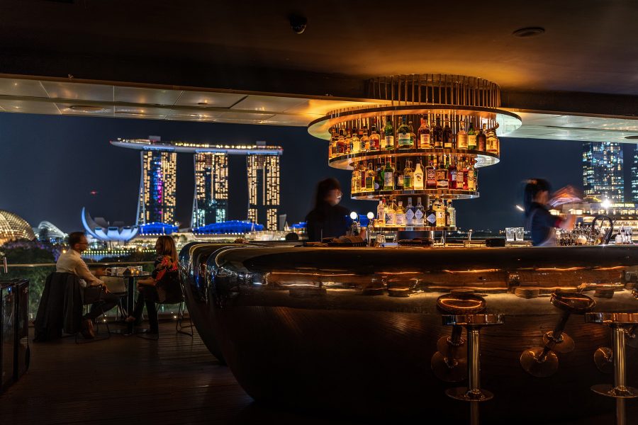 The interior of Smoke and Mirrors is dimly lit, allowing a spectacular nighttime view of the Marina Bay Sands in the distance, illuminated against the night sky. 