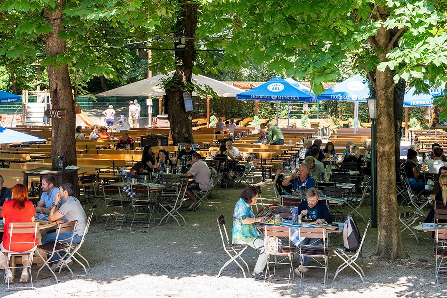 Groups of diners are seated outdoors under the shade of trees at beer gardens like the Hofbräukeller am Weinerplatz in Munich. 
