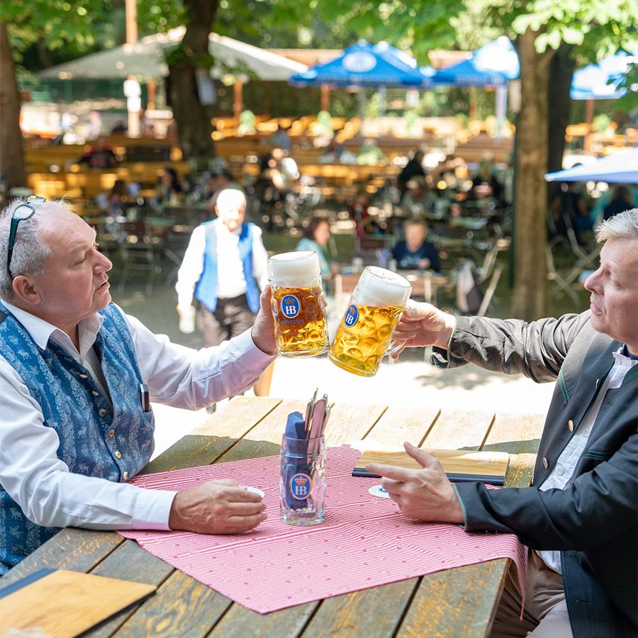 A table of two, seated outdoors in a Munich beer garden, clink their beer glasses. 
