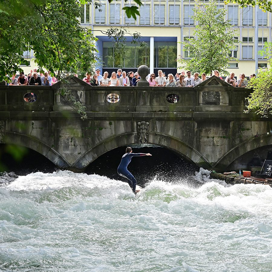A man surfs on Eisbach Canal in Munich as onlookers watch from a bridge above the water.
