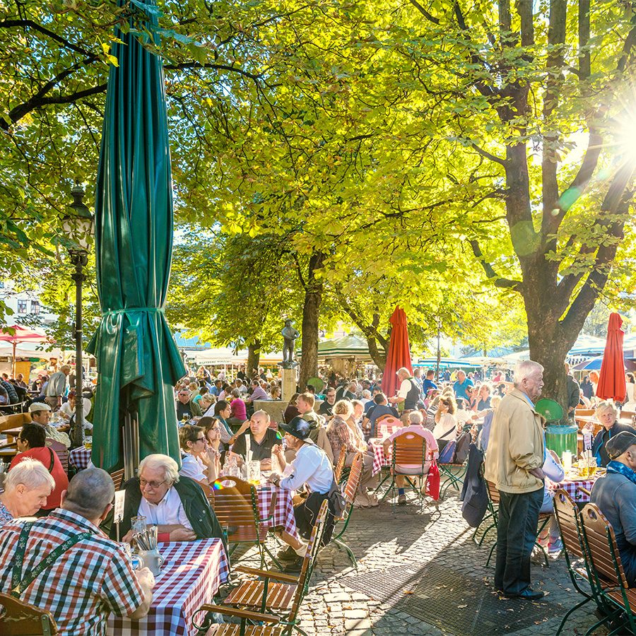 Groups of diners seated outdoors at Viktualienmarkt under the shade of trees on a sunny day. 
