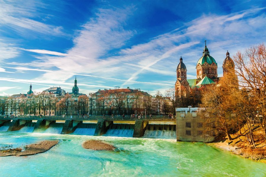 The water of the Isar appears turquoise on a sunny day in Munich, along a riverbank where a row of buildings is situated.