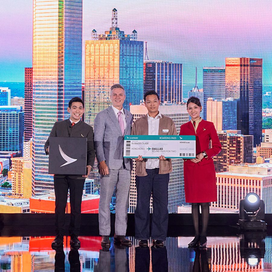 Two Cathay Pacific cabin crew, one male and one female, stand next to Cathay General Manager of Brand, Insights and Marketing Communications Edward Bell and a male winner of the prize draw in front of a backdrop showing the Dallas skyline at The Diamond Drinks in Hong Kong.