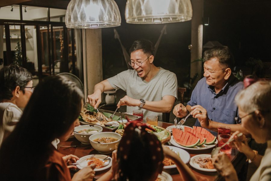 Man laughing as he serves food around a dining table surrounded by family.