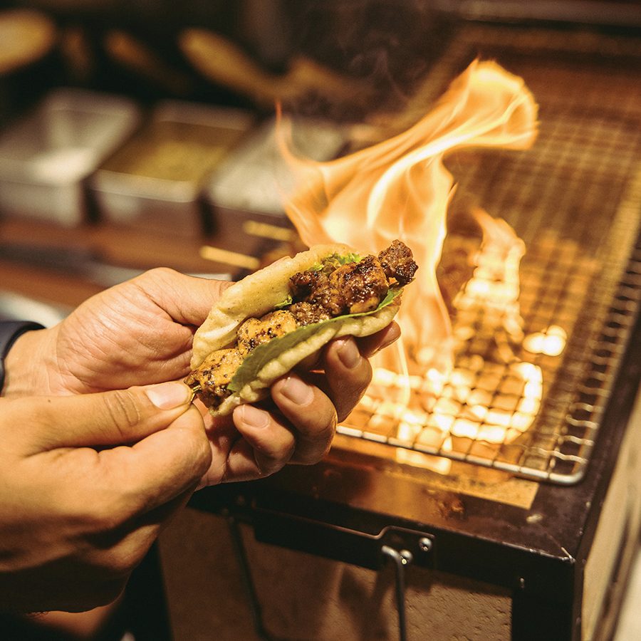 A close-up of a person holding a small kebab over a flaming grill, served at Papa’s restaurant in Mumbai.