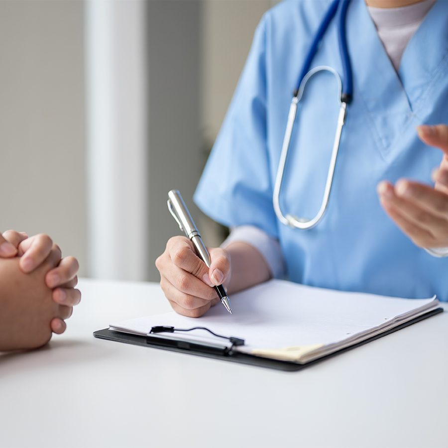  A patient’s clasped hands rest on a table as a medical professional with a pen and clipboard explains their diagnosis. 