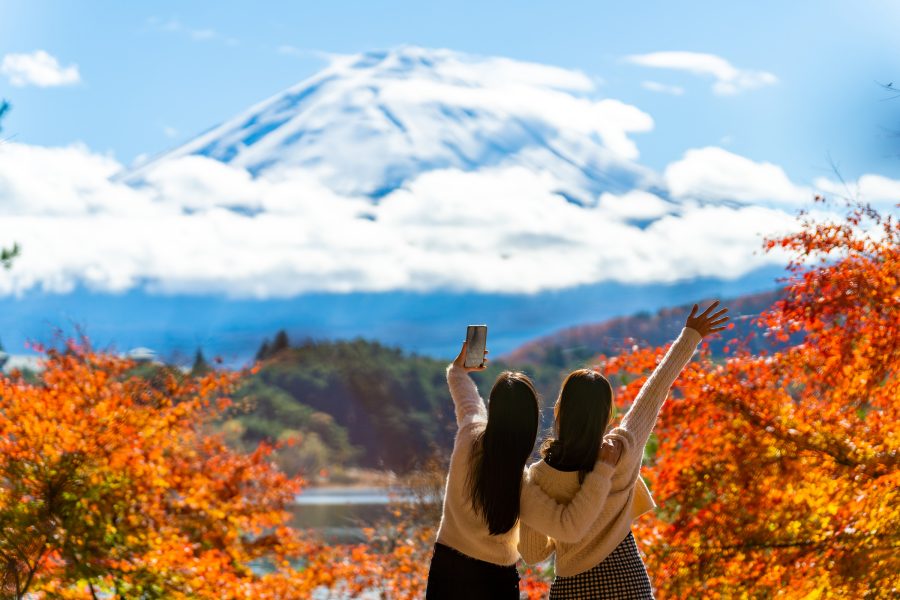 A shot of two women looking towards white clouds and Mount Fuji, surrounded by autumn leaves on a bright day, taken from behind