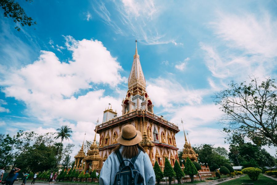 A shot of a woman wearing a hat looking towards Wat Chalong temple in Phuket on a sunny day, taken from behind // 