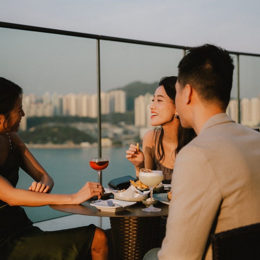 A man and two women enjoy cocktails at a rooftop bar with a seaview in Hong Kong