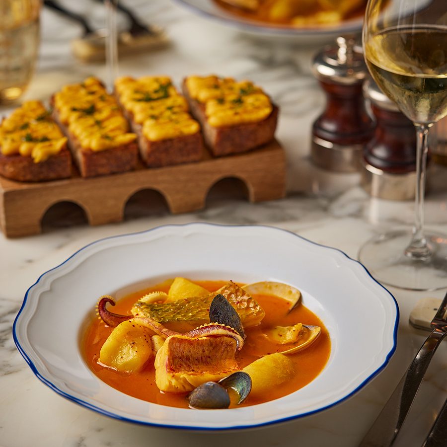 A close-up of a bowl of bouillabaisse on a marble dining table with bread and glasses of white wine, served at Marmo Bistro in Rosewood Hong Kong.