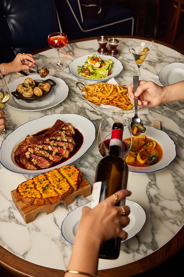 People eating and serving wine at a marble dining table, featuring a plate of côte de boeuf à la bordelaise and bouillabaisse, served at Marmo Bistro in Rosewood Hong Kong.