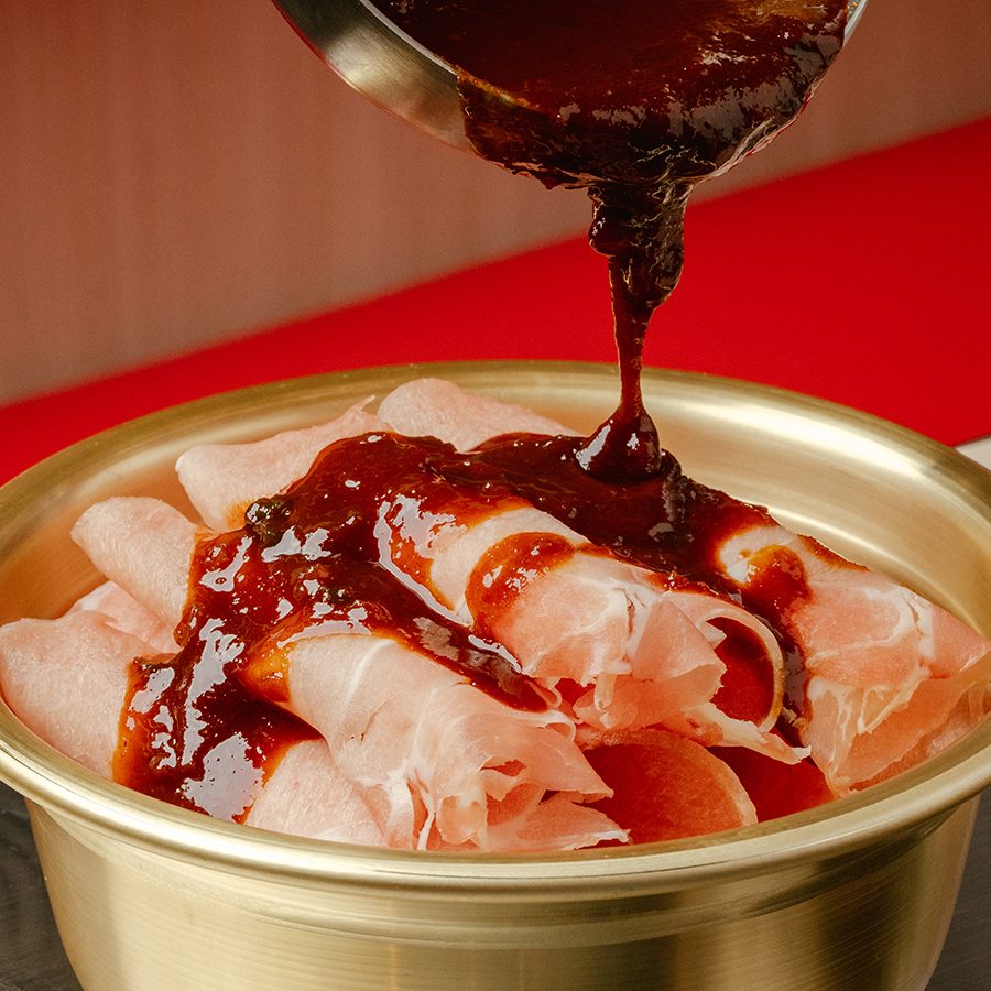 A close-up of a person pouring sauce over a bronze bowl of Yeoltan Bulgogi.