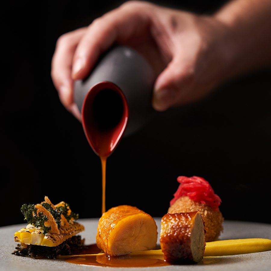 A close-up of a person pouring sauce over a dish of three yellow chicken, cromequis, neck boudin, kal, sweetcorn and smoked mayonaise, served at Té Bo in Hong Kong.