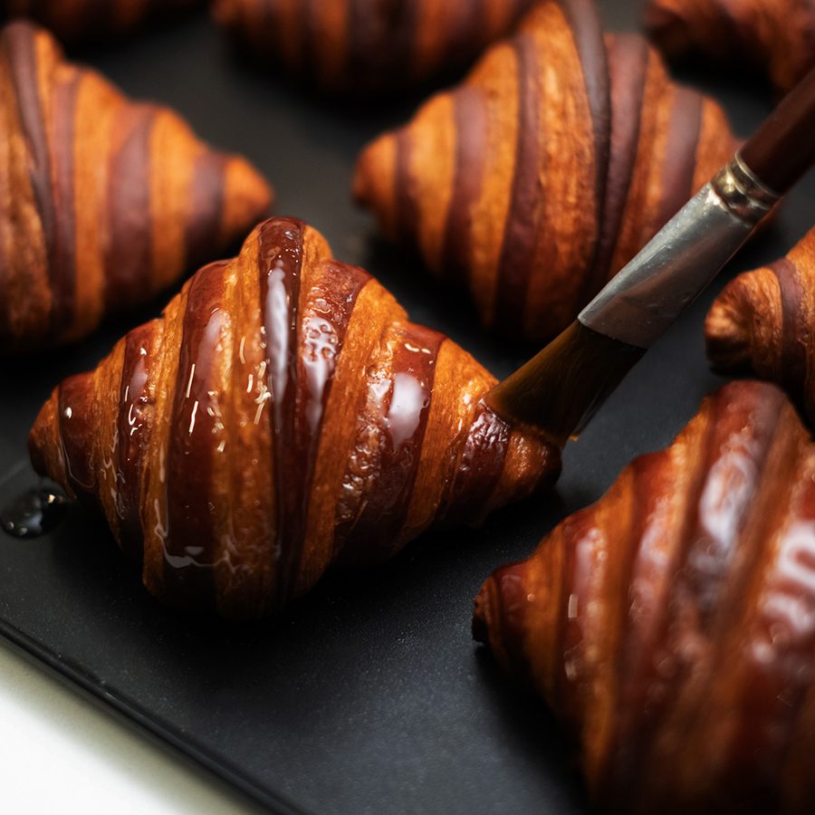 A close-up of a person brushing melted butter on a tray of mini chocolate croissants, served at Manam Chocolate Karkhana café in Hyderabad