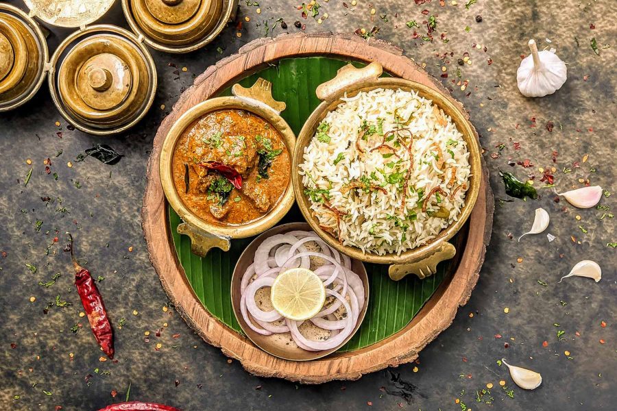 A top-down view of a mutton curry with plain pulao rice and a side dish of sliced red onion and lemon, served on a banana leaf and wooden board
