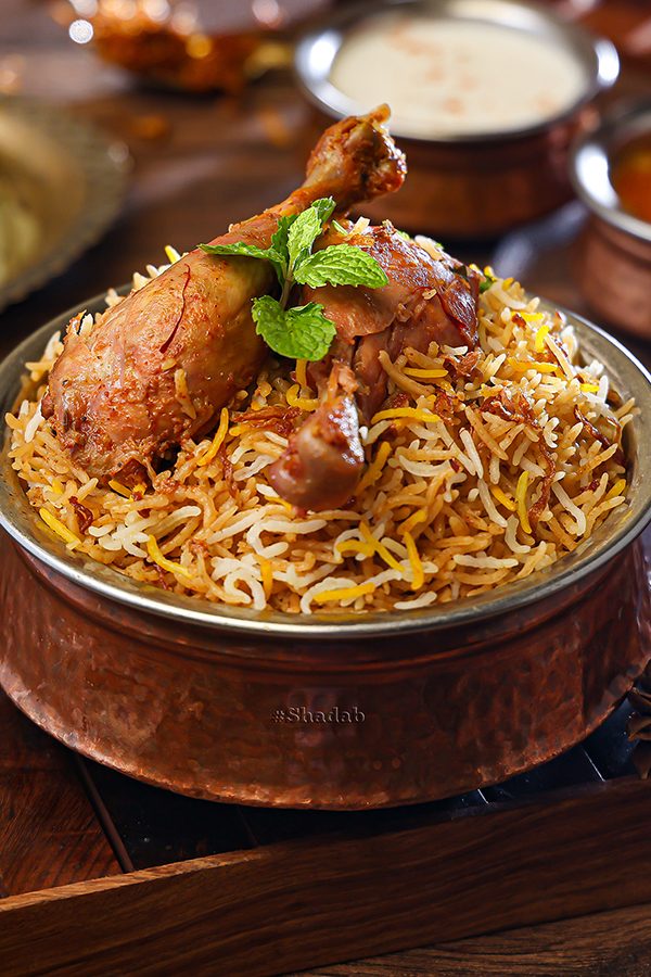 A close-up of a chicken biryani in a bronze bowl with raita and mirchi-ka-salan sauces in the background, served at Shadab in Old City in Hyderabad