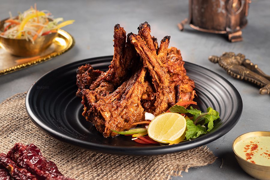 A close-up of a plate of curried tandoori lamb chops with sauces and vegetables in the background, served at Shadab in Old City in Hyderabad