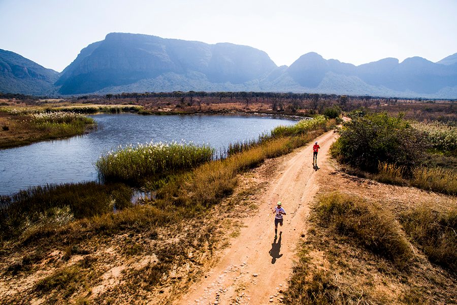A drone shot of two Big Five Marathon runners on the trail of the Entabeni Safari Conservancy in South Africa on a sunny day, with a view of grove, lake and mountains in the background