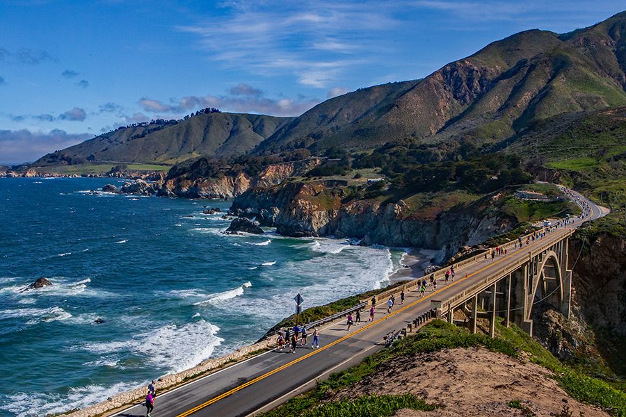 A bird’s eye view of international marathon runners running from a distance, on the bridge along the coast of the Big Sur in California during daytime with a view of the blue sky, sea and mountains in the background.