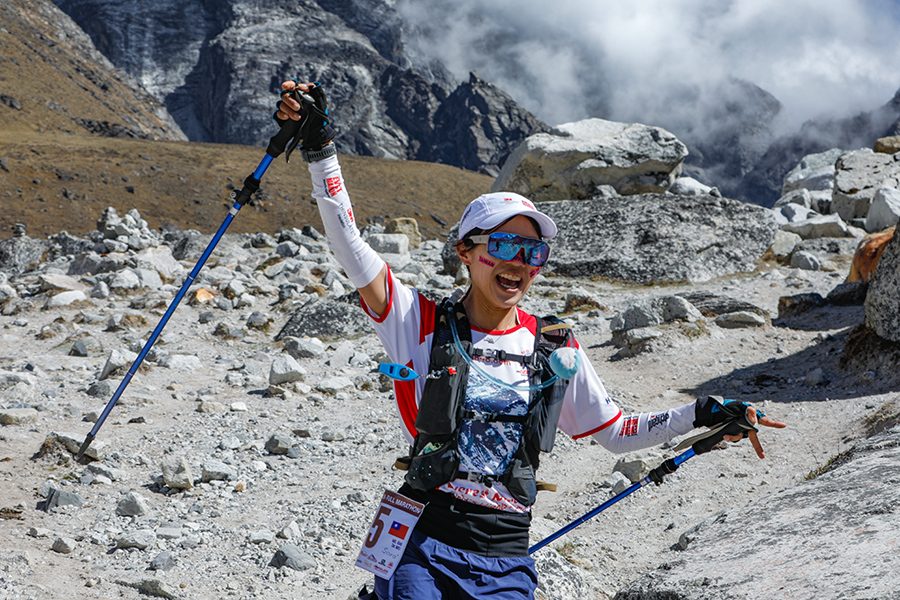 A woman trail runner cheering and smiling running down the rocky trail of the Everest Base Camp for the Everest Marathon, with clouds and rocky mountains in the background