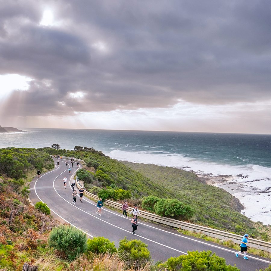 A drone shot of marathon runners on the open road for the Great Ocean Road Marathon in Australia, with views of the vast ocean and sun shining through cloudy skies. 